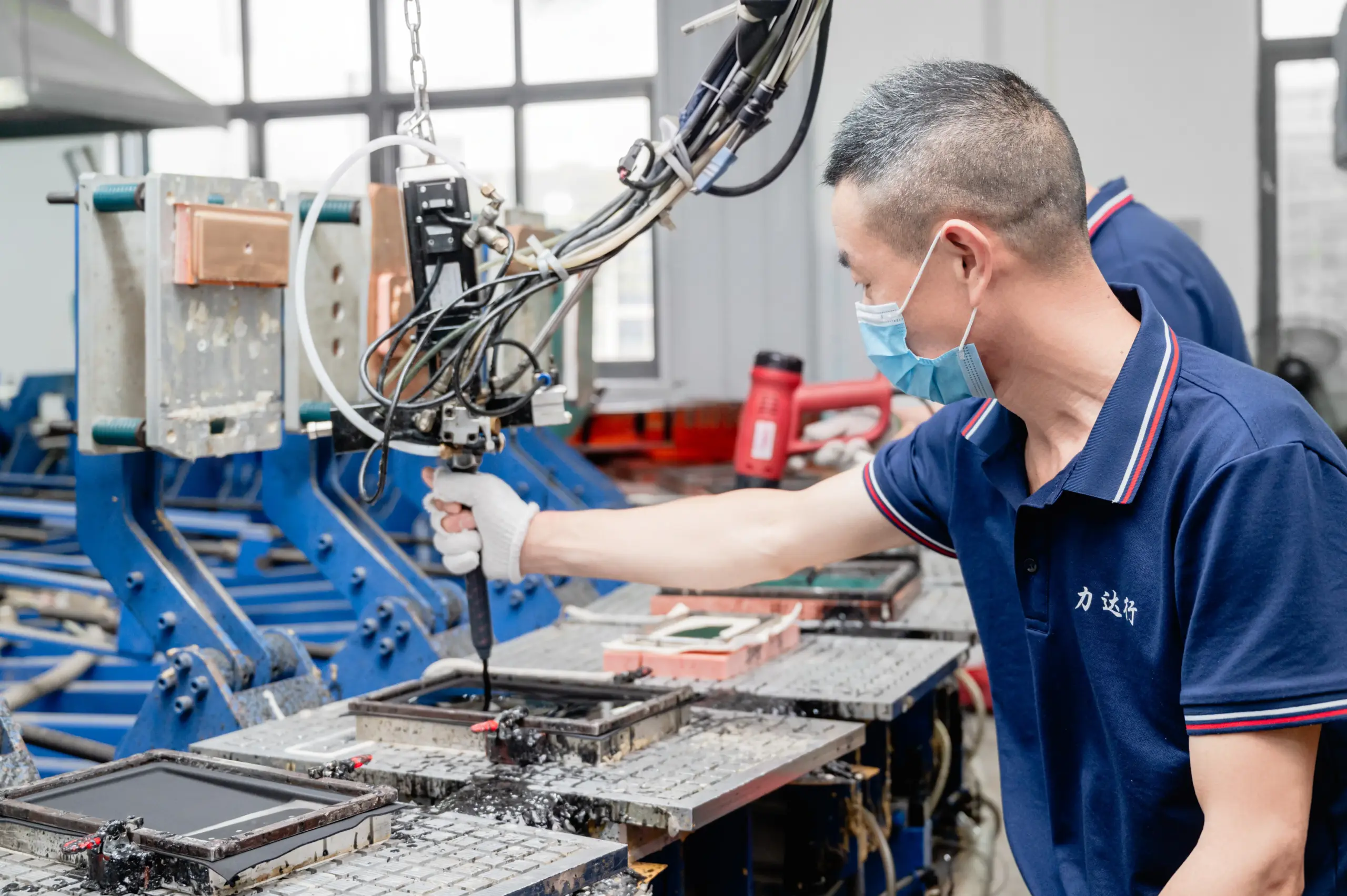 A factory worker in a blue uniform and mask applies sealant to manufacturing molds using an automated dispensing tool.