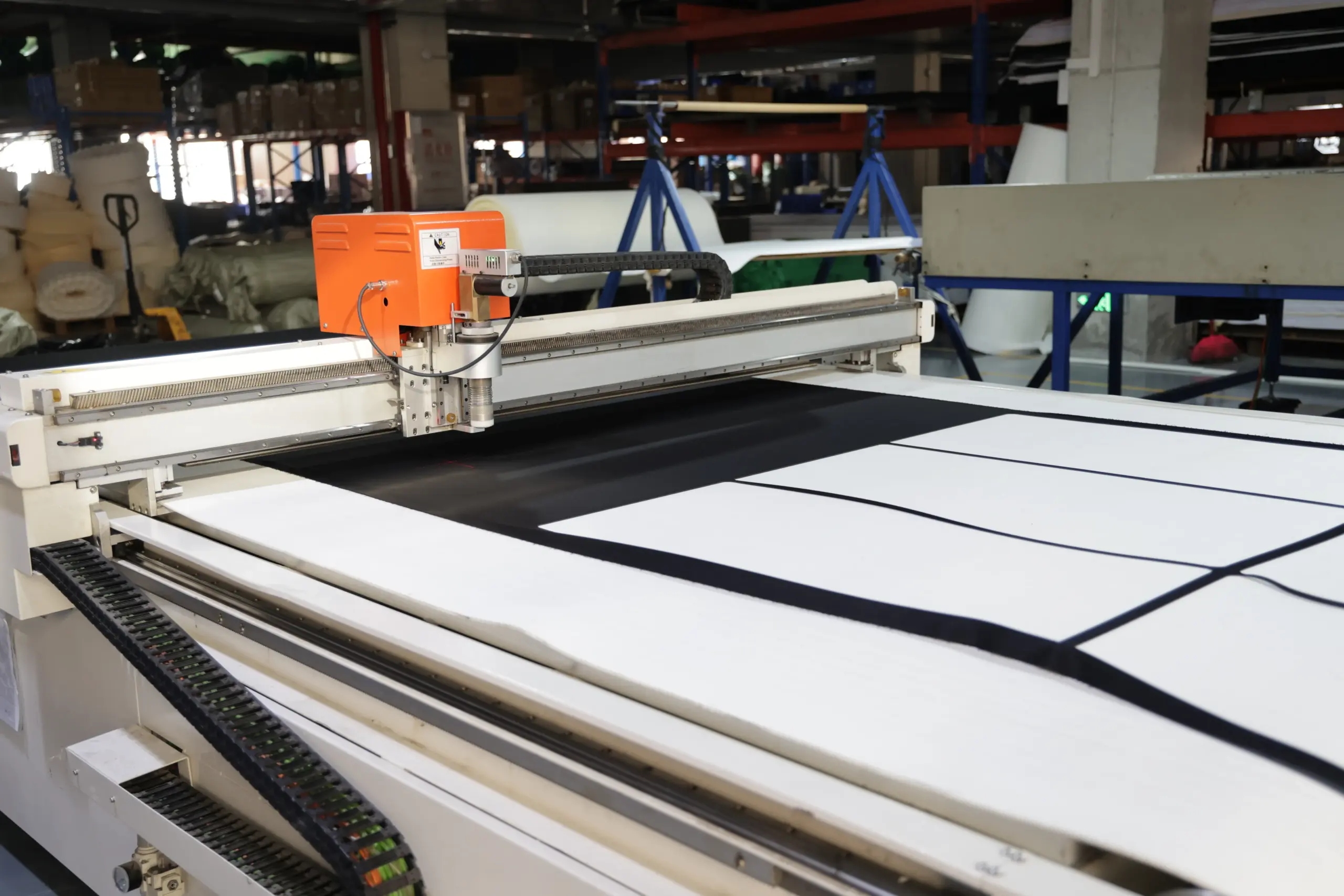 An industrial CNC cutting machine with an orange head precisely cutting white material on a large flatbed table in a factory.